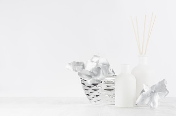 Elegant home decor with glass white aroma diffuser bottles, sticks, silver branch, bowl on white wood table, wall.