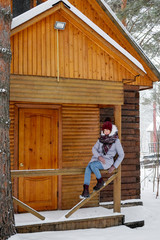 Woman sitting on snowy porch wooden rustic cottage in the winter woods. Portrait of a girl in a warm gray coat and burgundy knit scarf.