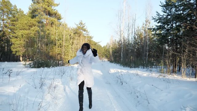 smiling African-American woman in white fur coat runs along snowy path and laughs in winter pine-tree forest slow motion