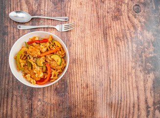  Flat lay of a white bowl of a spicy chicken pasta dinner with a stainless-steel fork and spoon on a wooden background with copy space