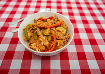 Selective focus on the contents of a spicy chicken and pasta dish with a stainless-steel fork and spoon on a red and white checked table cloth