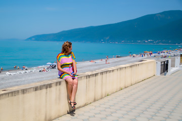 A woman sits on a fence by the sea in a bright multicolored dress