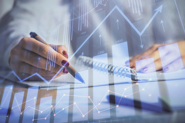 Double exposure of woman's hands making notes with forex graph hologram. Concept stock market...