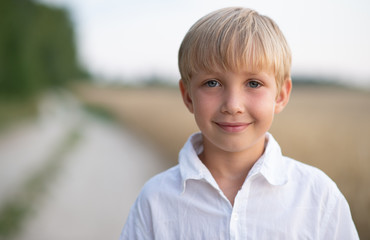 Face close - up of a boy of eight years. Portrait of a smiling boy in nature looking at the camera. Happy blond boy with a smile on his face. Child in a white shirt with blue eyes in nature.