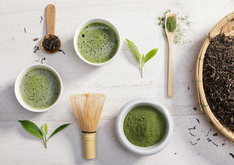 Organic matcha green tea powder in bowl with wire whisk and green tea leaf on white stone table,...