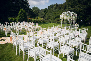 Elegant wedding luxury ceremony outdoors. An arch decorated with orchids surrounded by white wooden chairs on a green lawn strewn with rose petals.