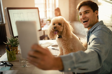 Happy freelance worker using touchpad while taking selfie with his dog in the office.