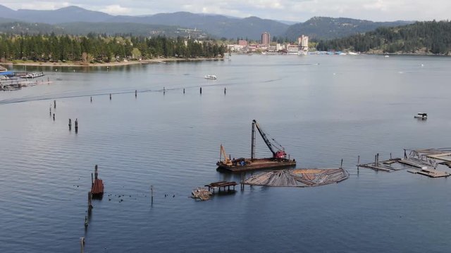 An aerial shot circling around a barge holding an excavator and a crane in the middle of Lake Coeur d'Alene with Coeur d'Alene, the resort and Tubbs Hill in the background.