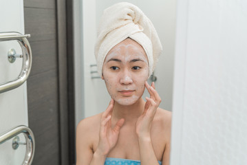 Asian girl in a mask with a towel in front of the mirror in the bathroom. the concept of self-care, moisturizing and cleansing the skin. care of youth and beauty