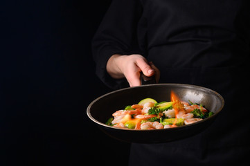 Chef cooks shrimps with vegetables, in a pan with fire. Frying and cooking. Oriental cuisine. Culinary, restaurant business, healthy and tasty food. Banner, on black background.