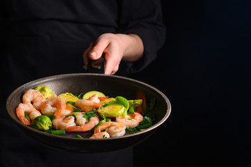 Chef cooks shrimps with vegetables, in a pan with fire. Frying and cooking. Oriental cuisine. Culinary, restaurant business, healthy and tasty food. Banner, on black background.