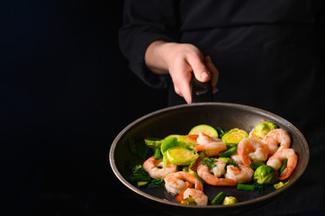 Professional chef cook on a dark background prepares seafood, shrimp with fire and vegetables. Roasting and cooking veggie food. Black background, Space for design.