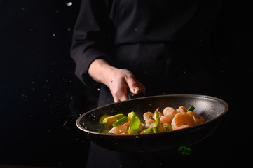 Chef cooks with fire in a pan shrimp with vegetables. On a black background, restaurant service