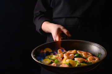 Professional chef cook on a dark background prepares seafood, shrimp with fire and vegetables. Roasting and cooking veggie food. Black background, Space for design.