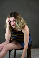 Portrait of a pretty student girl with beautiful curly hair in front of the camera on a gray background. Concept vertical photo of a young woman in a black T-shirt and blue skirt sitting on a chair.