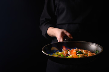 Chef cooks shrimps with vegetables, in a pan with fire. Frying and cooking. Oriental cuisine. Culinary, restaurant business, healthy and tasty food. Banner, on black background.