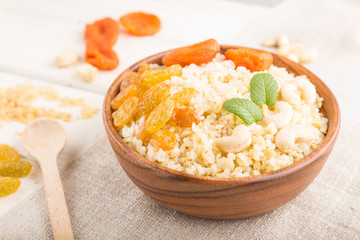 Bulgur porridge with dried apricots,  raisins and cashew in wooden bowl on a white wooden background and linen textile. Side view, selective focus.