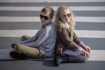 girl child curly blonde in a gray pleated skirt and boy, brown jacket and sunglasses sitting on the pavement zebra crosswalk