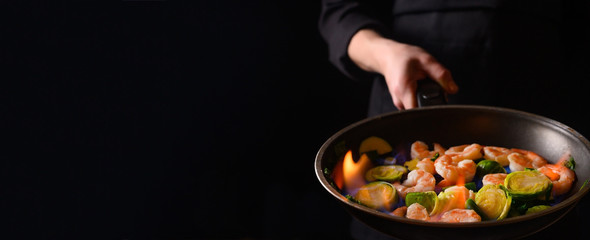 Chef cooks shrimps with vegetables, in a pan with fire. Frying and cooking. Oriental cuisine. Culinary, restaurant business, healthy and tasty food. Banner, on black background.
