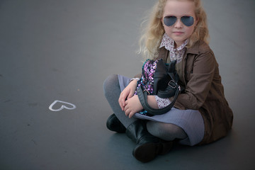 girl child curly blonde in a gray pleated skirt, brown jacket and sunglasses sitting on the pavement