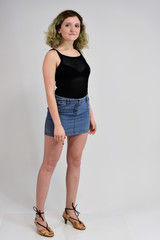Fototapeta premium A horizontal photo concept of a young woman in a black t-shirt and blue skirt is standing in front of the camera on a white background. Full-length portrait of a pretty girl with beautiful curly hair.