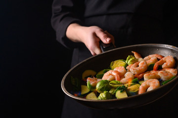 Chef cooks shrimps with vegetables, in a pan with fire. Frying and cooking. Oriental cuisine. Culinary, restaurant business, healthy and tasty food. Banner, on black background.
