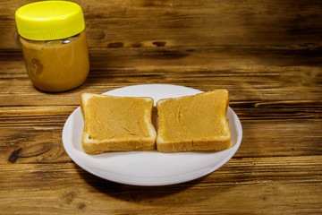 Jar of peanut butter and plate with two sandwiches with peanut butter on wooden table