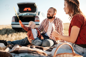 Happy Young Family Mom and Dad with Their Little Son Enjoying Summer Weekend Picnic on the Car Outside the City, Playing with Bubbles