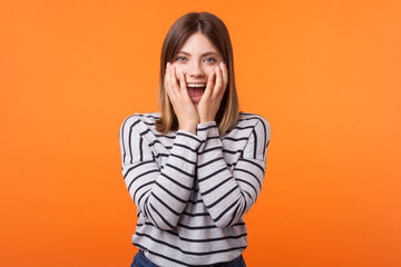 Portrait of shocked young woman with brown hair in long sleeve striped shirt standing, looking with amazement and hiding face in arms, unexpected news. indoor studio shot isolated on orange background