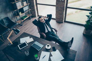 High angle above view photo of handsome business guy drink coffee relaxing break legs on table looking notebook screen wear specs black blazer shirt pants tie suit sit chair office indoors