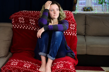 Fototapeta premium Portrait of a girl with beautiful magnificent hair rejoices on a sofa. Horizontal concept photo of a young woman in a sweater and jeans sits on a chair in front of the camera in the home interior.