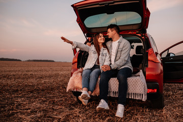Naklejka na ściany i meble Young Happy Couple Dressed Alike in White Shirt and Jeans Sitting at Their New Car Trunk, Beautiful Sunset on the Field, Vacation and Travel Concept