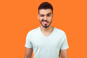 Obraz premium Portrait of playful handsome brunette man with beard in casual white t-shirt standing with one eye closed, winking and flirting at camera with grin. indoor studio shot isolated on orange background