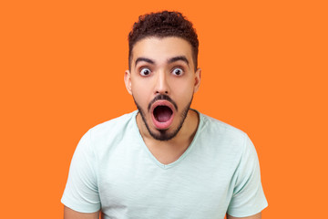 I can't believe this. Portrait of emotional brunette man with beard in white t-shirt staring at camera with big eyes and open mouth, shocked by news. indoor studio shot isolated on orange background