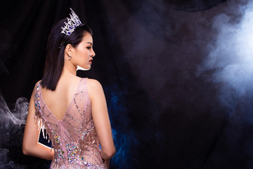 Portrait of Miss Pageant Beauty Contest in Pink sequin Evening Ball Gown with Diamond Crown, Asian Woman points waves hand to side over dark background smoke copy space © Jade