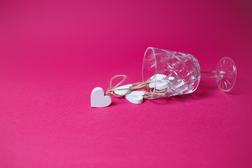 Small white wooden hearts in the lying wine glass on pink background with copy space.