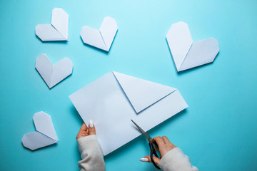 Many white origami hearts on blue background with sheet of paper and scissors in the center. Saint Valentine's day card with woman making origami heart on blue background.