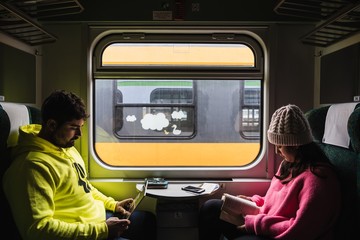 Couple sitting across from each other in front of a train window entertaining each other with a tablet and a book