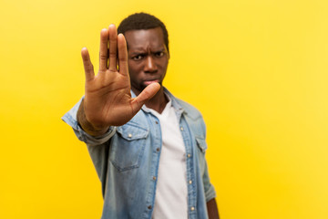 Prohibition. Portrait of serious angry man in denim casual shirt frowning showing stop gesture with palm of the hand, saying no, warning expression. indoor studio shot isolated on yellow background