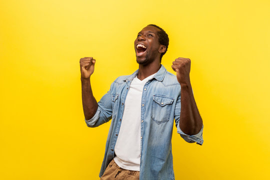 Portrait of ecstatic overjoyed handsome man in denim casual shirt expressing winning gesture with raised fists and screaming, celebrating victory. indoor studio shot isolated on yellow background