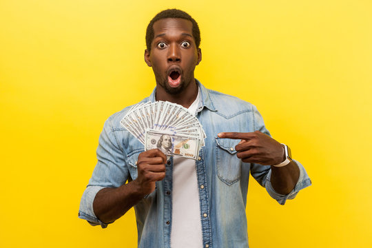 Portrait of amazed young man in denim casual shirt pointing at dollar bills and looking shocked by large amount of money, unexpected bonus, cashback. indoor studio shot isolated on yellow background