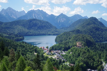 A bird's eye view of the valley with the bluest lake, small village and landmark Hohenschwangau Castle. The lake is surrounded by heavily forested mountains. It's a beautiful summer day.