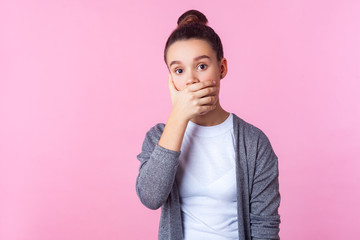 I won't tell! Portrait of scared brunette teenage girl with bun hairstyle in casual clothes covering mouth with hand, looking frightened intimidated. indoor studio shot isolated on pink background