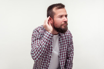 Portrait of deaf bearded man in plaid shirt standing with hand near ear and listening carefully to...