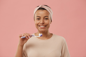 Head and shoulders portrait of beautiful mixed race woman holding toothbrush and smiling at camera in morning while standing against dust pink background, copy space