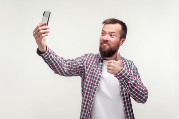 Like this phone! Portrait of excited handsome bearded man in plaid shirt taking selfie or talking on video call and showing thumbs up, satisfied with mobile service. studio shot, white background