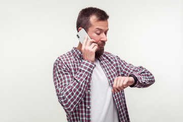 Portrait of punctual bearded man in plaid shirt talking on phone and checking time on wristwatch, dissatisfied with delay, deadline, mobile communication. indoor studio shot isolated, white background