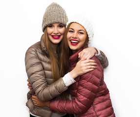 Close up fashion portrait of Two cheerful beautiful girls friends posing for camera inside. Bright makeup, hats and winter casual style. White background, not isolated.Having fun together.