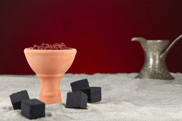 Shisha in a clay bowl, prepared for Smoking a hookah, stands on the sand surrounded by coals and a metal graceful cezve, decorated with Arabic arnament on an maroon background.