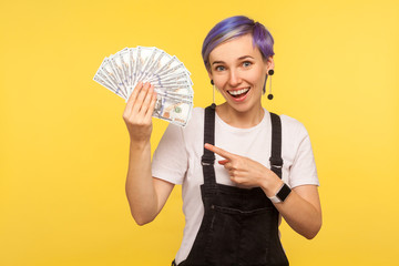 Portrait of joyful trendy hipster girl with violet short hair in denim overalls pointing at fan of dollar banknotes and looking at camera with toothy smile. isolated on yellow background, studio shot